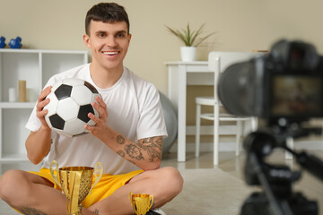 Sporty happy male blogger with gold cups, medals and soccer ball recording video at home