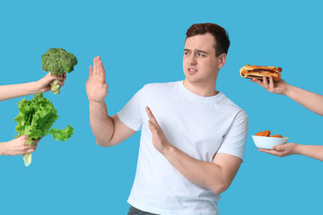 Young overweight man rejecting healthy food for unhealthy on blue background. Weight gain concept