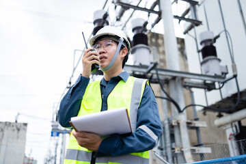 Engineer is working at high voltage power substation performing inspection and check of infrastructure using walkie talkie and clipboard for safety and accuracy