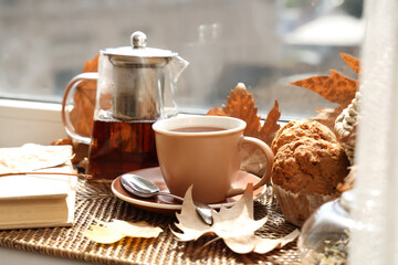 Teapot with cup of hot beverage, tasty cupcakes and autumn leaves on windowsill