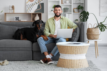 Young man with Dobermann dog using laptop on sofa at home