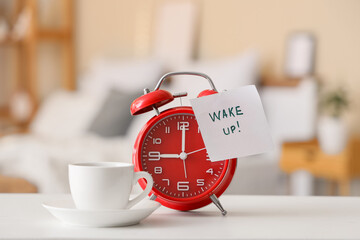 Alarm clock, cup of coffee and sticky note with text WAKE UP on bedside table in bedroom, closeup