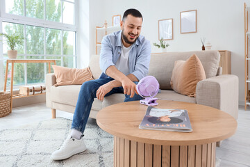 Young man turning mini fan on table at home