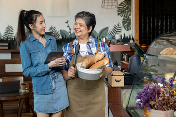 Asian senior woman in apron holding basket of bread smiling and talking with young female staff in denim dress using tablet. Friendly atmosphere in small family-run bakery with teamwork
