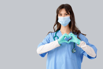 Female medical intern in mask making heart gesture on light background