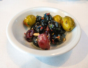 A small white dish holds a colorful mix of marinated olives—green, black, and purple—glistening with oil and herbs, accented by crushed garlic and red pepper flakes. A savory Mediterranean bite.