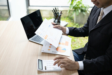 A man in a suit is sitting at a desk with a laptop and a calculator. He is holding a piece of paper and he is working on some financial documents. Concept of professionalism and focus
