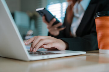 A businesswoman in a suit uses her smartphone at a desk with documents, notebooks, and a takeaway coffee cup.
