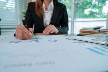 A businesswoman in a suit types on a laptop at a desk covered with charts, graphs, and business documents.