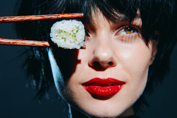 Beautiful woman with short hair holds sushi with chopsticks, showcasing vibrant colors and a striking expression against a dark background.
