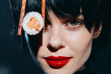 Close-up portrait of a woman with dark hair, red lips, and sushi, showcasing elegance and curiosity against a dark backdrop.