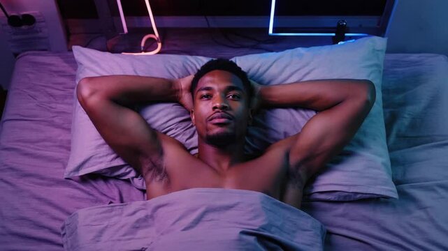 shirtless ripped man lying on bed, covered partially by a grey  blanket, with his head resting on two pillows