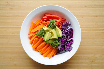 Overhead View of a White Bowl Filled with Sliced Carrots Red Bell Peppers Avocado and Shredded Red Cabbage on a Light Brown Wooden Surface
