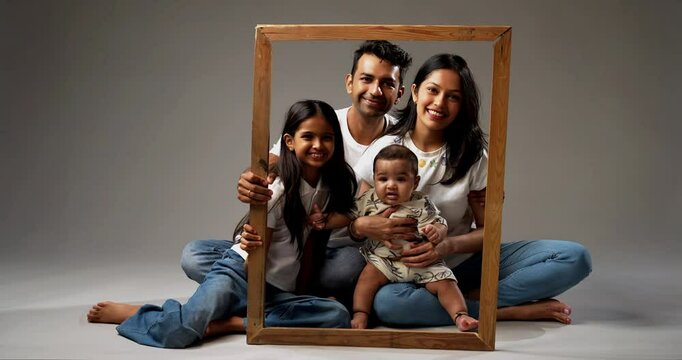 Family looking through photo frame while smiling at camera, Indian parents with daughter and infant baby posing happily together, sitting indoors against gray background, showing unity and love