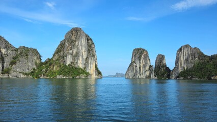 Naklejka premium Towering limestone karsts rise from the calm emerald waters of Ha Long Bay, Vietnam, under a clear blue sky.