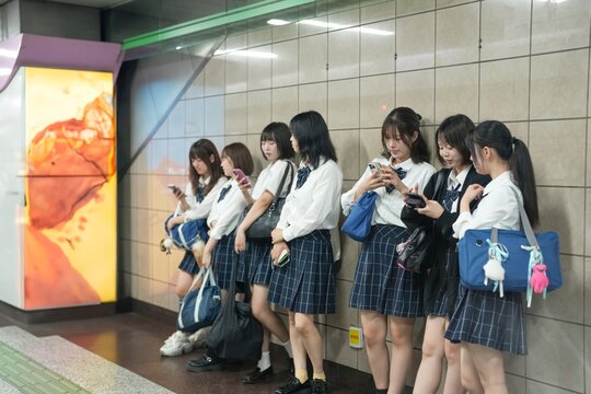 On a spring morning in Shanghai, Japanese high school girls in uniforms sit on a subway platform wall, chatting and laughing while waiting for their train during a quiet commute in a foreign city.