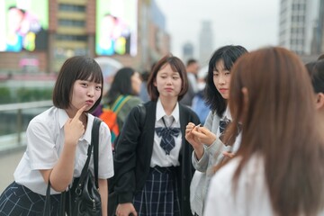 In early summer in Shanghai, near The Bund, Japanese high school girls in uniforms chat and laugh in a busy tourist area, their cheerful presence adding youthful charm to the city's historic vibe.