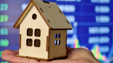Wooden house model in hand against a screen with financial market data, representing housing market investment and trends - Powered by Adobe
