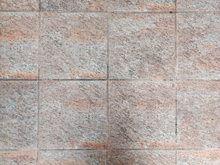 A tiled surface displays square tiles with a rough, granular texture and a mottled brown and beige appearance, separated by dark grout lines, conveying durability and a rustic or earthy aesthetic