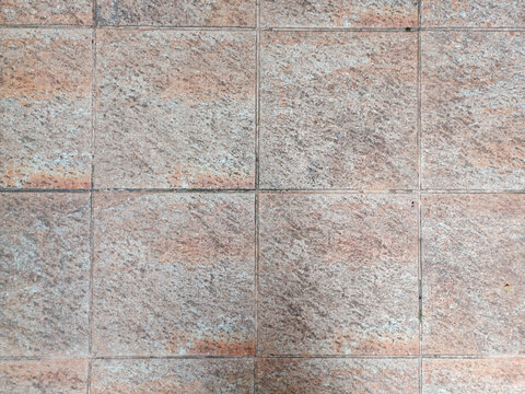 A tiled surface displays square tiles with a rough, granular texture and a mottled brown and beige appearance, separated by dark grout lines, conveying durability and a rustic or earthy aesthetic