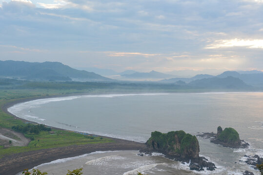 Stunning panoramic view of Teluk Love in Jember, Indonesia, featuring its distinctive heart-shaped bay, dark sandy beach, and distant misty mountains under the soft light of an early morning sky
