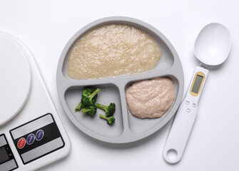 Baby food divider plate with healthy food and kitchen scales, spoon on white background. Top view