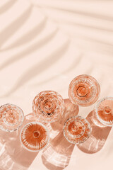Minimal summer party concept, set glasses of rose wine or champagne on pink with palm leaf shadows and light reflections. Bright shiny wineglass with sparkling alcohol drink at sunlight, top view