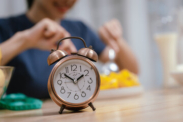 An antique-style alarm clock sits on a table, with a person eating in the blurred background,...