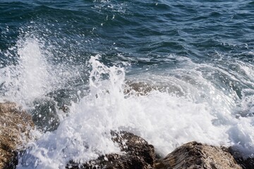 Fototapeta premium Rocky shore with turquoise sea water rolling in waves and creating splashes and thick white sea foam.