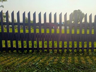 Obraz premium A wooden picket fence in a grassy field under a clear sky. The sun casts shadows on the ground, creating a serene outdoor atmosphere.