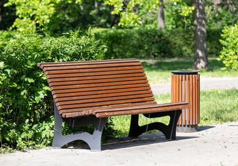 A wooden bench sits in a park next to a trash can