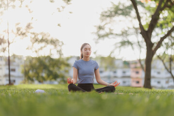 Fototapeta premium A young woman in workout attire practices yoga in a peaceful pose at a public park. The soft evening sunlight filters beautifully through the trees, creating a calm and serene atmosphere.