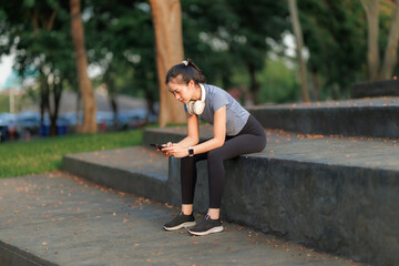 A young woman takes a break after exercising, relaxing with a cheerful smile. She wears headphones, listens to music, and chats with her friend on the phone, enjoying the peaceful moment.