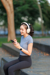 A young woman takes a break after exercising, relaxing with a cheerful smile. She wears headphones, listens to music, and chats with her friend on the phone, enjoying the peaceful moment.