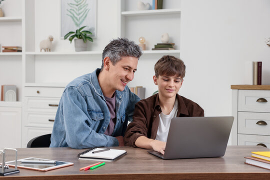 Smiling father and his son doing homework with laptop at table indoors