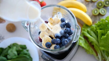 Chef is pouring milk into a blender containing bananas, blueberries, and other fruits, surrounded by fresh ingredients like spinach, bananas, kiwi, and lettuce, preparing a healthy smoothie