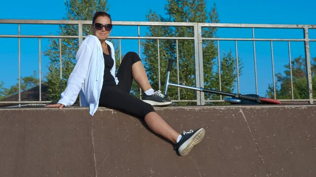 Young woman posing sitting on a skatepark ledge. Young woman in sportswear poses confidently while sitting on the ledge of an urban skatepark ramp, showcasing her scooter under a blue sky