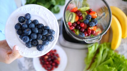 Woman holding a plate of fresh blueberries over a blender filled with kiwi, strawberries, cranberries, and blueberries, with bananas and lettuce nearby for a healthy smoothie