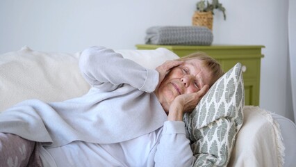 Elderly woman experiencing a severe headache, lying on a comfortable sofa with her hands on her temples, seeking relief from the pain in a cozy home environment