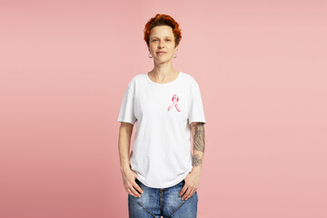 Confident non binary person wearing pink ribbon for breast cancer awareness posing with hands in...