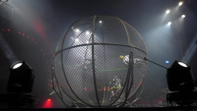 Three acrobats ride motorcycles in a large metal round cage in a circus with colorful lighting. Performance of circus performers on a motorcycle. Dangerous motorcycle stunts. Acrobats on motorcycles.