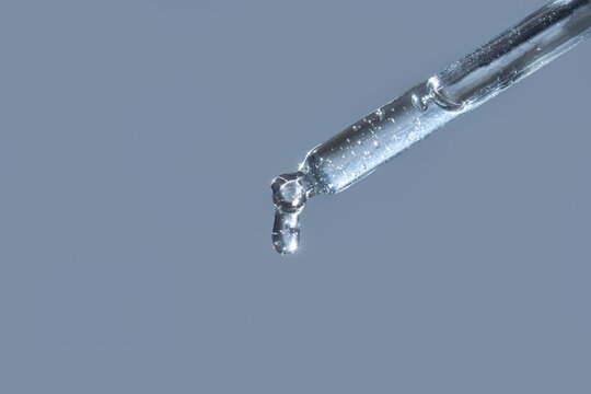 Close-up of transparent liquid drop from glass pipette against light blue background.