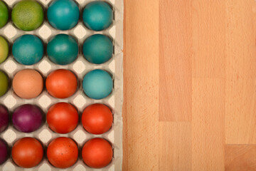 A top-down shot shows a carton filled with colorful Easter eggs, including shades of green, blue, peach, purple, and red, on a wooden surface