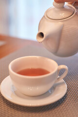 White teapot pouring freshly brewed tea into white cup on table with soft lighting.