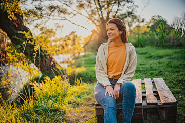 Woman sitting on wooden bench enjoying sunset at a riverside park surrounded by greenery in...