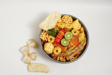 Top view of a bowl of spicy Indonesian seblak with chicken feet, tofu, crackers, sausage, and lime, served on a white background.