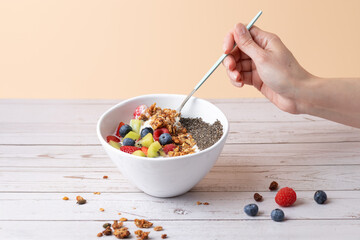 Front view of female hand holding a spoon inside a breakfast bowl with kefir, fruit, granola and chia. Peach background and rustic surface. Copy space. Concept of balanced eating and natural living