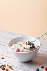 Spoon inside a bowl of kefir with fresh fruit, granola, and chia seeds. Peach-colored background and white wooden table. Copy space. Concept of natural breakfast and healthy lifestyle.