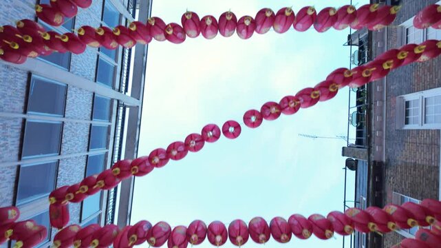Chinese lanterns hanging between urban buildings, gently swaying in wind against pale blue sky, representing traditional asian festival decoration and cultural celebration