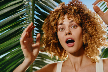 surprised woman with curly hair captured outdoors under green palm leaves, wearing white tank top, expressing amazement and natural beauty in bright sunlight.